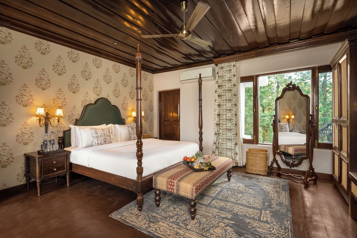 A four-poster bed with a wooden headboard, a bedside table with lamps, a bench with a tray of fruit, and a large window with a view of greenery.