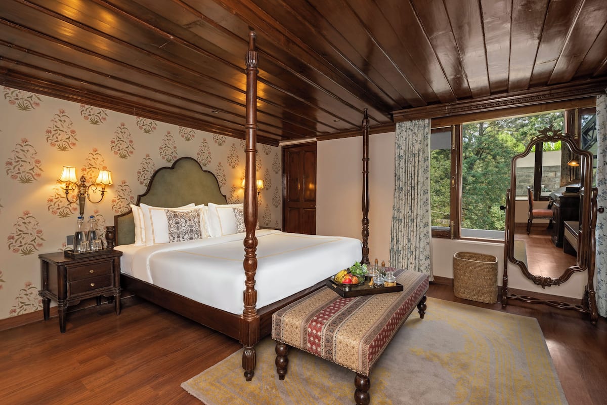 A four-poster bed with a wooden headboard, a bedside table with lamps, a bench, and a window with a view of greenery.
