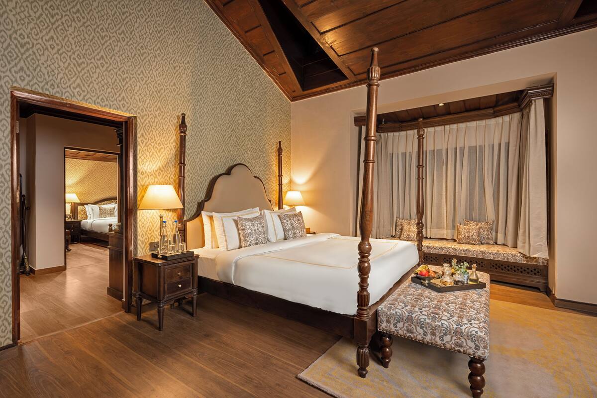 A four-poster bed with a footboard, a bedside table with a lamp, a bench with a tray, and a window with curtains.
