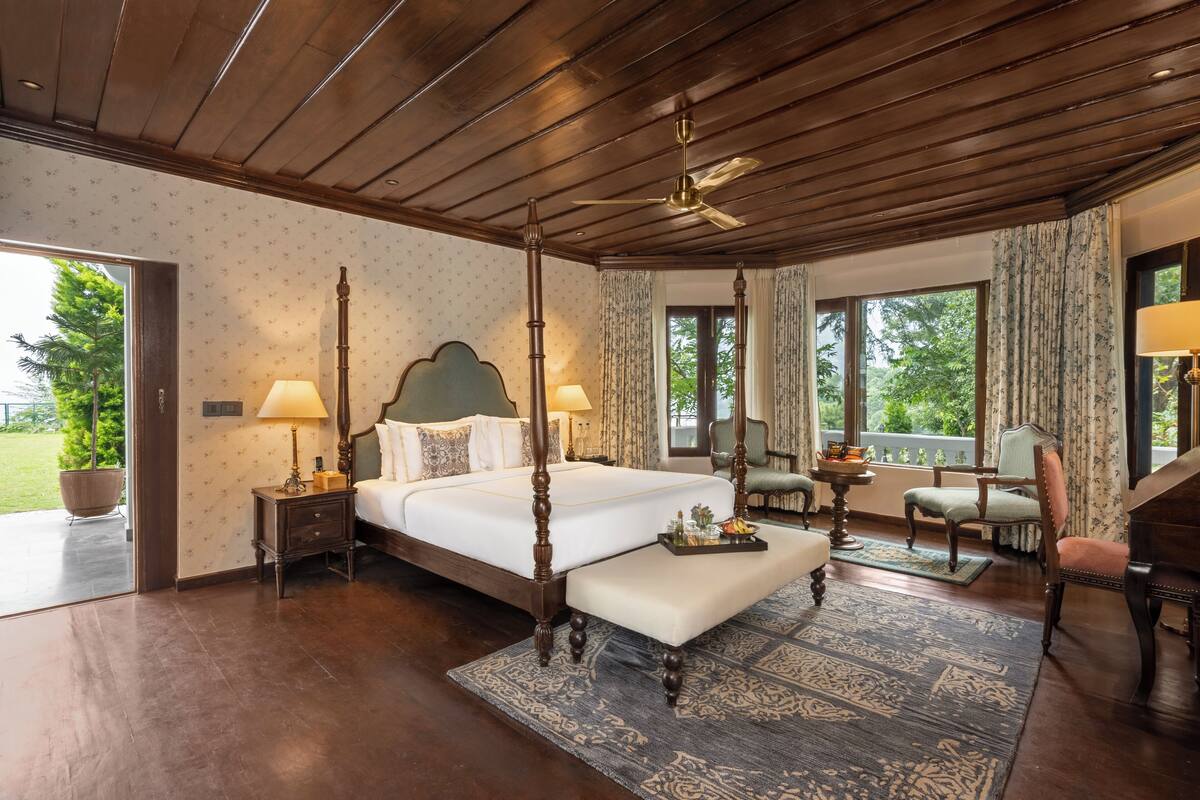 A four-poster bed with a wooden headboard, a sitting bench, a desk with a chair, and a view of the outdoors through large windows.