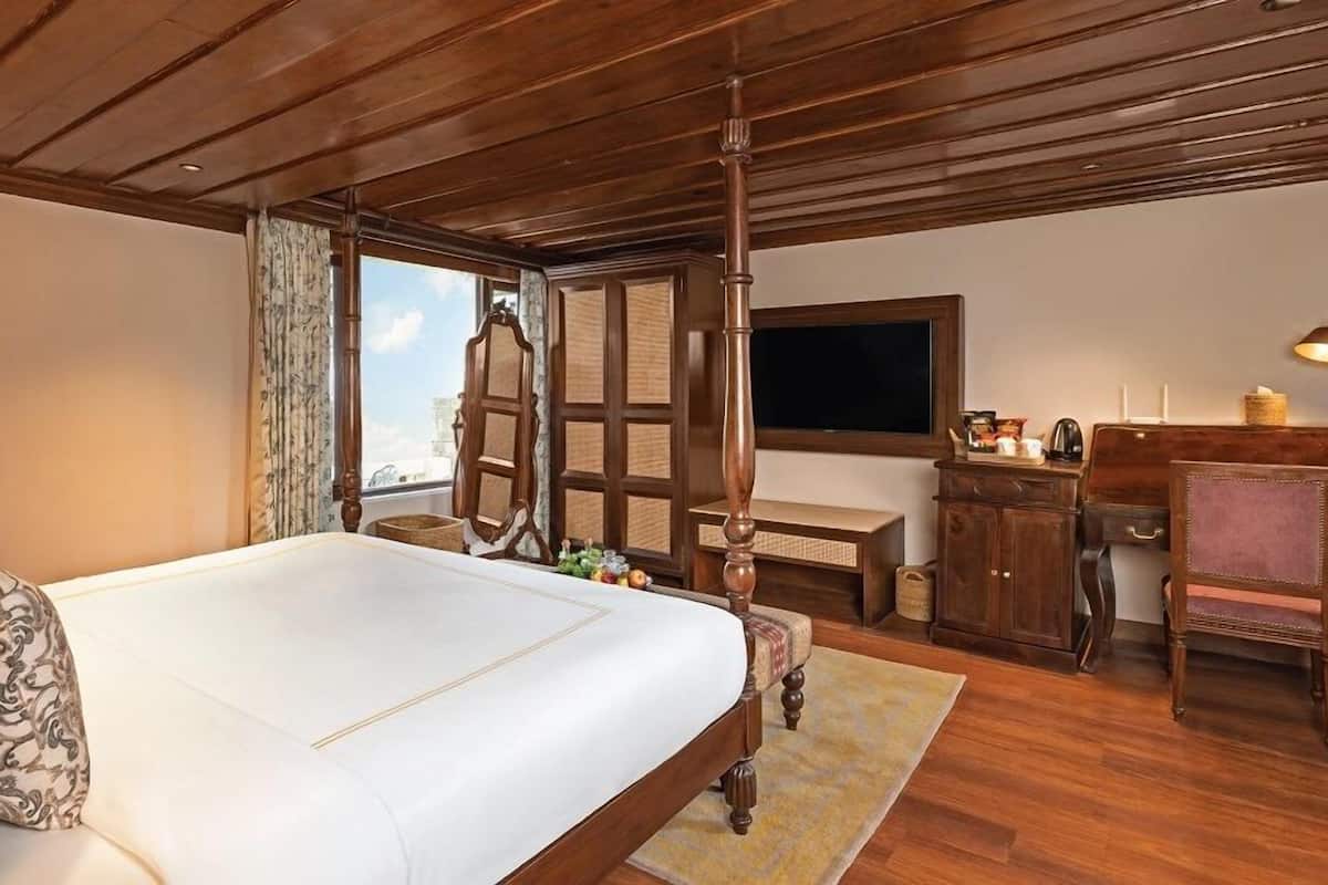 A bedroom with a large bed, wooden ceiling, a desk with a chair, a television, and a window with curtains.