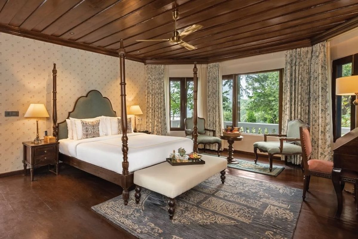 A four-poster bed with a wooden headboard, a bedside table with a lamp, a chair, and a view of greenery outside.