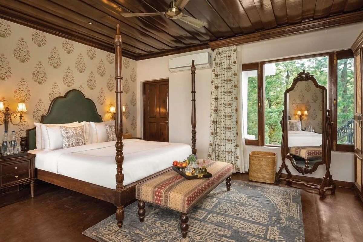A four-poster bed with a wooden headboard, a bedside table with a lamp, a bench with a tray, and a view of the outdoors through a door.