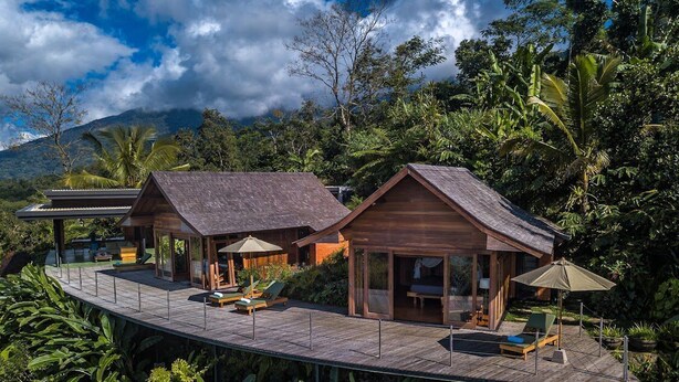 Una cabaña de madera con terraza, rodeada de exuberante vegetación y un telón de fondo montañoso.