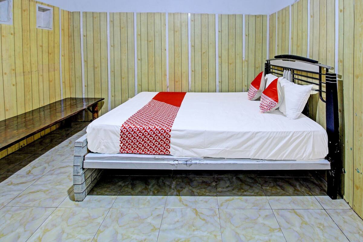 Una cama individual con una colcha blanca y roja estampada, un cabecero de madera y un banco a la izquierda.