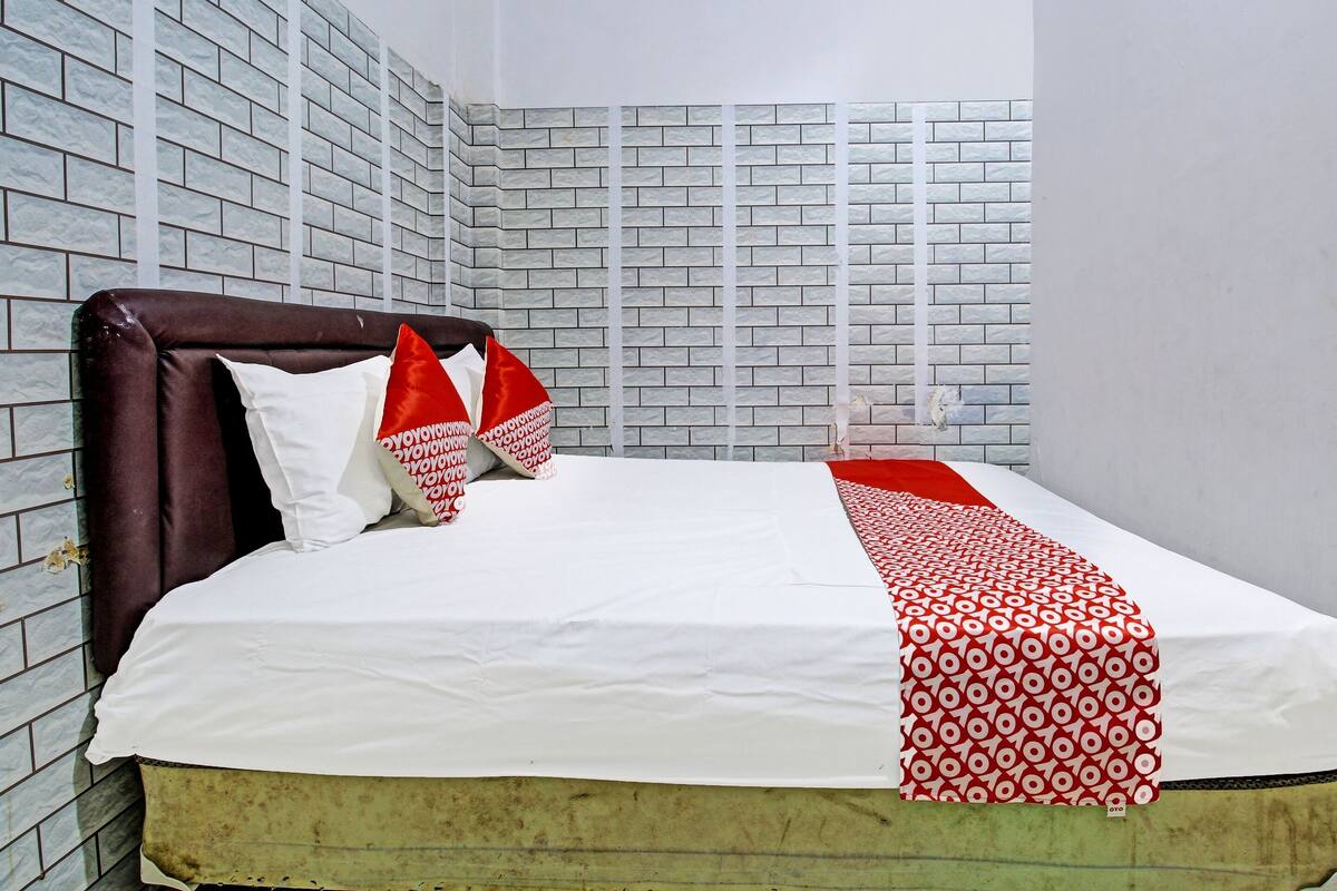 Una cama con colchón blanco, cabecera marrón y almohadas estampadas en rojo y blanco. La habitación tiene una pared con diseño de ladrillo y una cortina blanca.