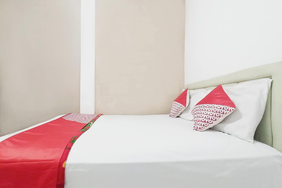 Cama com roupa de cama branca e almofadas vermelhas, cómoda de madeira e uma parede lisa.