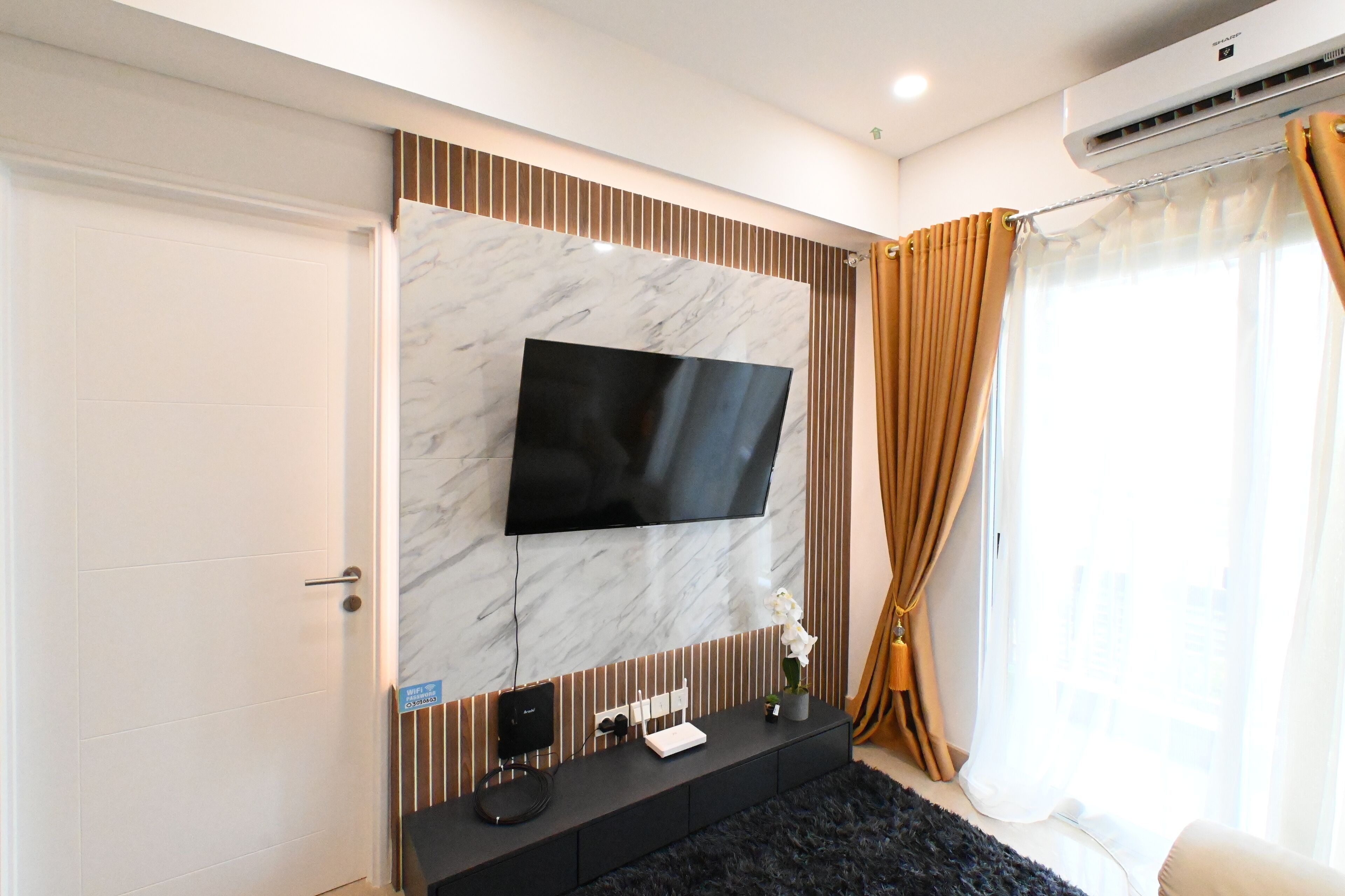 Habitación de hotel moderna con un televisor de pantalla plana grande montado en una pared de mármol, una mesa consola negra con un altavoz y una planta pequeña, y una ventana con cortinas color beige.