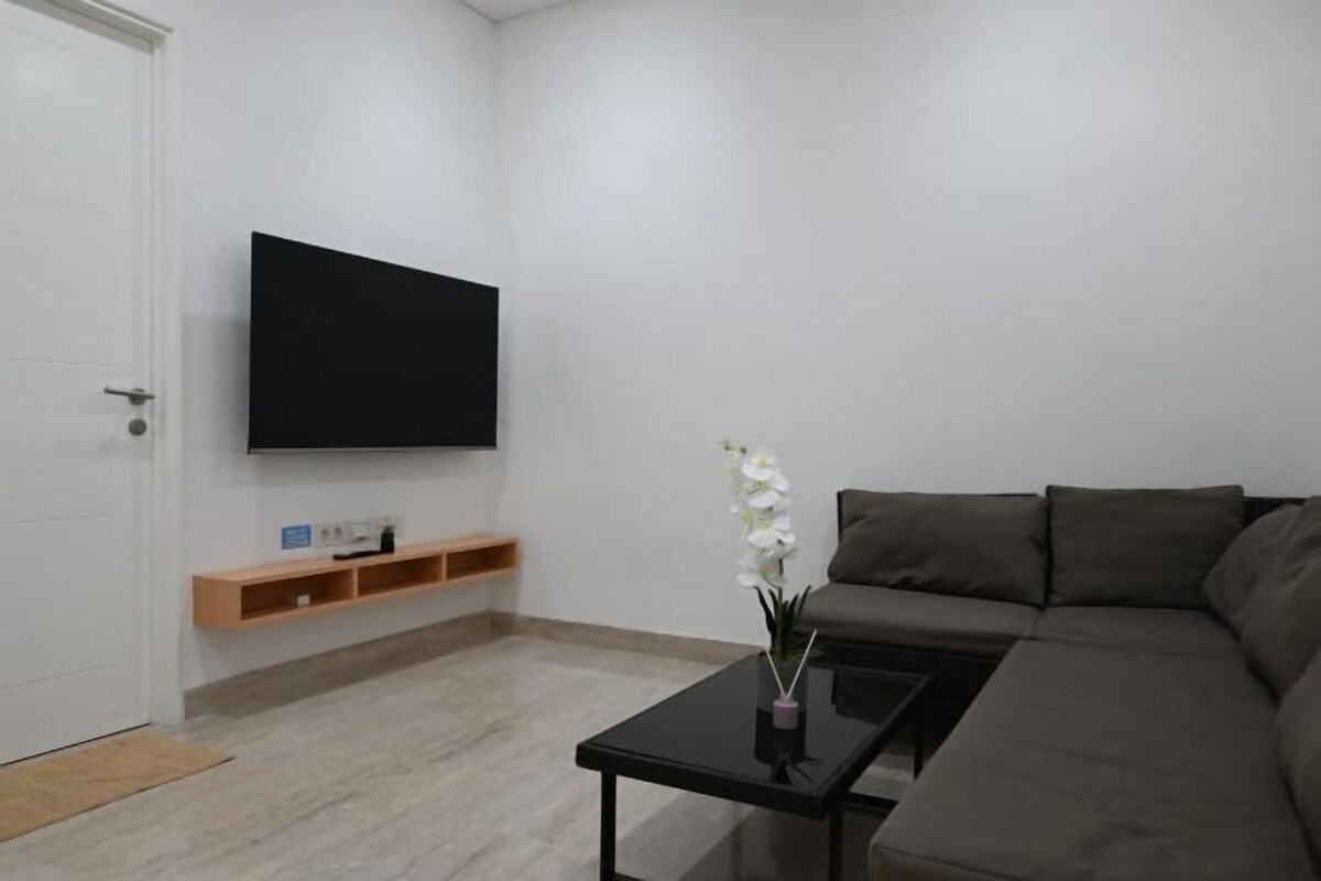 Una sala de estar moderna con un televisor de pantalla plana montado en la pared, un sofá de color oscuro, una mesa de centro y un pequeño estante de madera con algunos objetos.