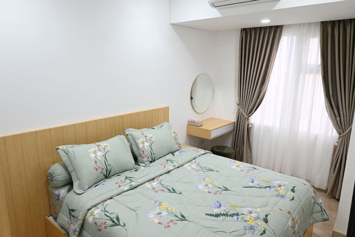 Ein Schlafzimmer mit Blumenmuster-Bettdecke, einem Holzkopfteil, einem Nachttisch mit Lampe und einem Fenster mit Vorhängen.