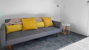 A grey sofa with two mustard yellow cushions, a small round side table, and a framed picture of animals on the wall. - Coastal Sanctuary - Brand New Modern Townhouse (Christchurch)