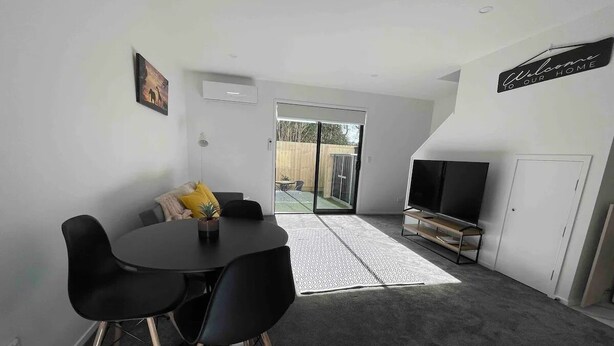 A modern living room with a dining area, a flat-screen TV, and a sliding glass door leading to a balcony. - Coastal Sanctuary - Brand New Modern Townhouse (Christchurch)