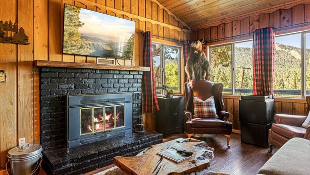 Ein gemütliches Blockhaus-Interieur mit Kamin, Holzwänden und einem großen Fenster mit Blick auf einen bewaldeten Bereich.