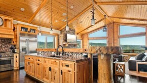 A spacious kitchen with wooden cabinets, a large island, and a stone backsplash. There is a stone fireplace and a large window overlooking a scenic view.