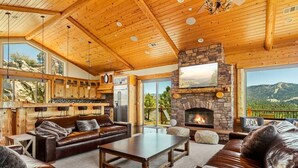 A spacious living area with a stone fireplace, wooden ceiling, and large windows offering a scenic view.