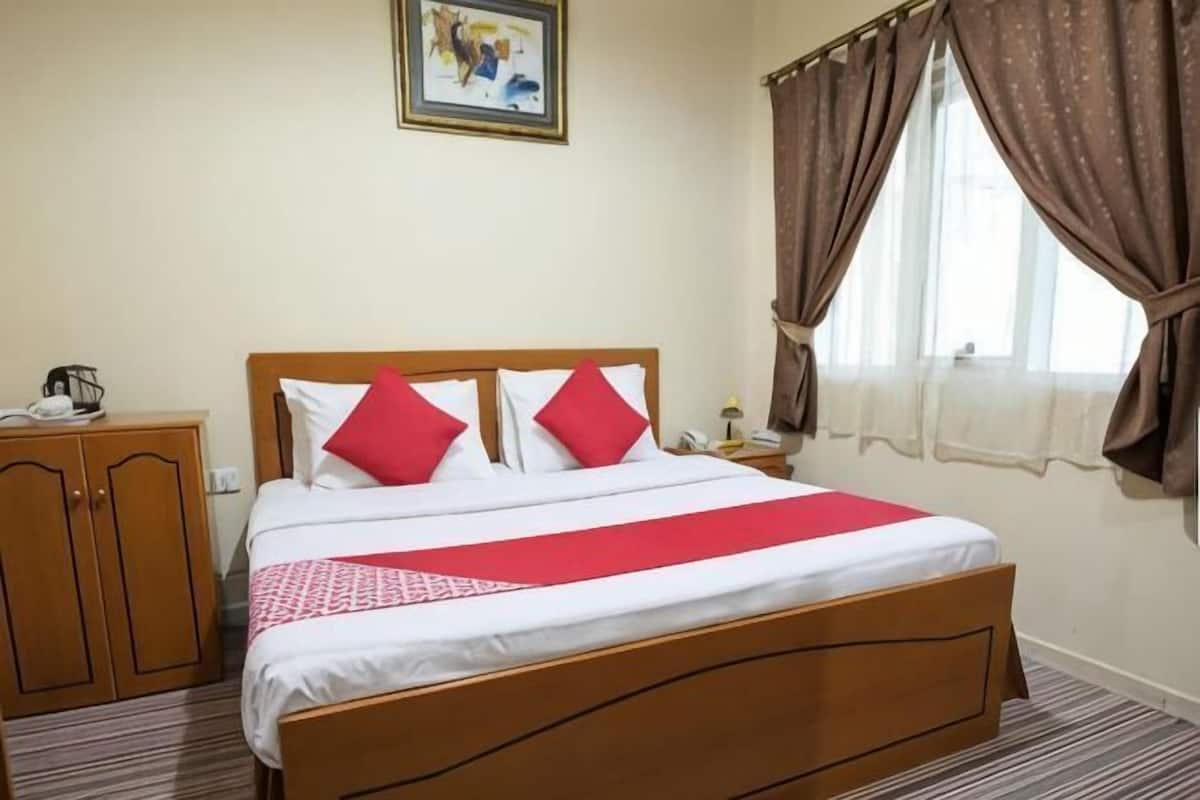 Une chambre d’hôtel avec un lit en bois, deux oreillers, une table de chevet, un téléphone, une fenêtre avec des rideaux et un tableau encadré au mur.