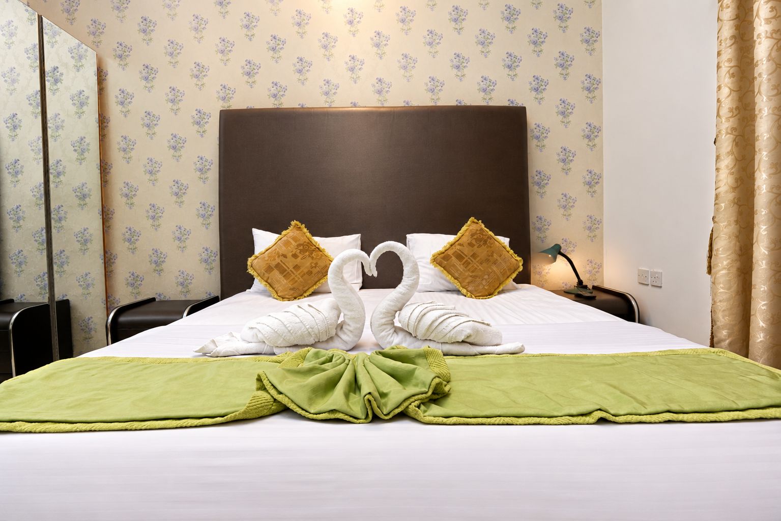 A hotel room with a neatly made bed, two pillows, a green throw blanket, and a headboard.