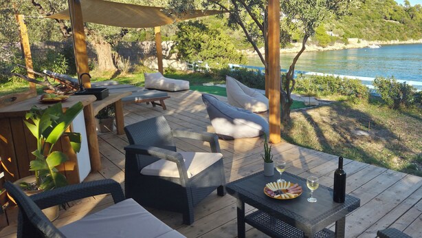 A wooden deck with outdoor furniture, a table with a wine bottle and glasses, and a scenic view of a lake and trees. - Actaea, Relaxing Villa by the Seashore (Alonissos)