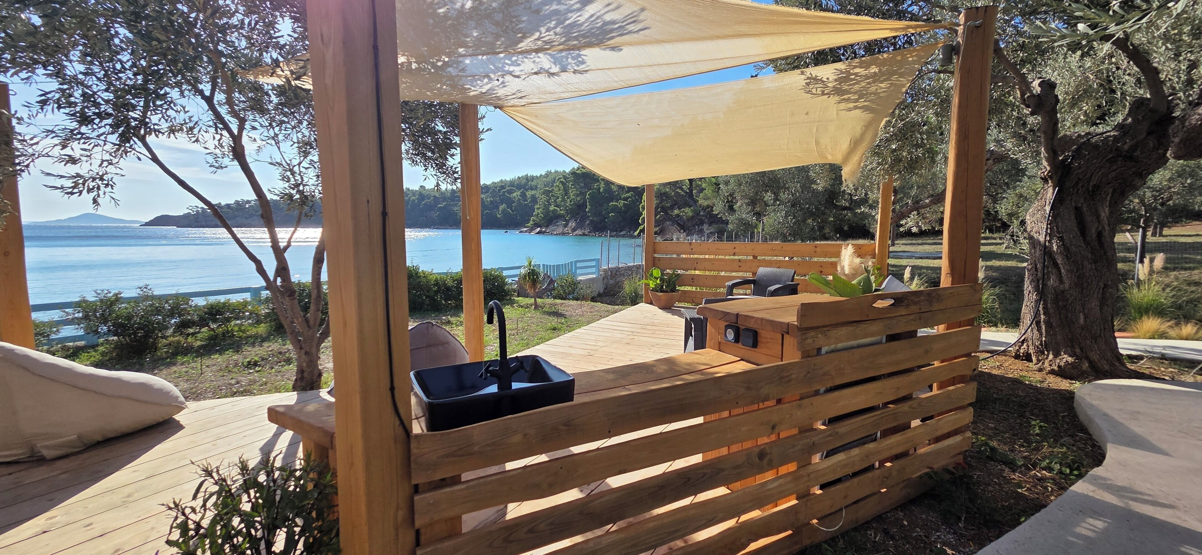 Eine Holzterrasse mit einer Bank, einem Waschbecken und einem schattigen Bereich mit Blick auf ein Gewässer.