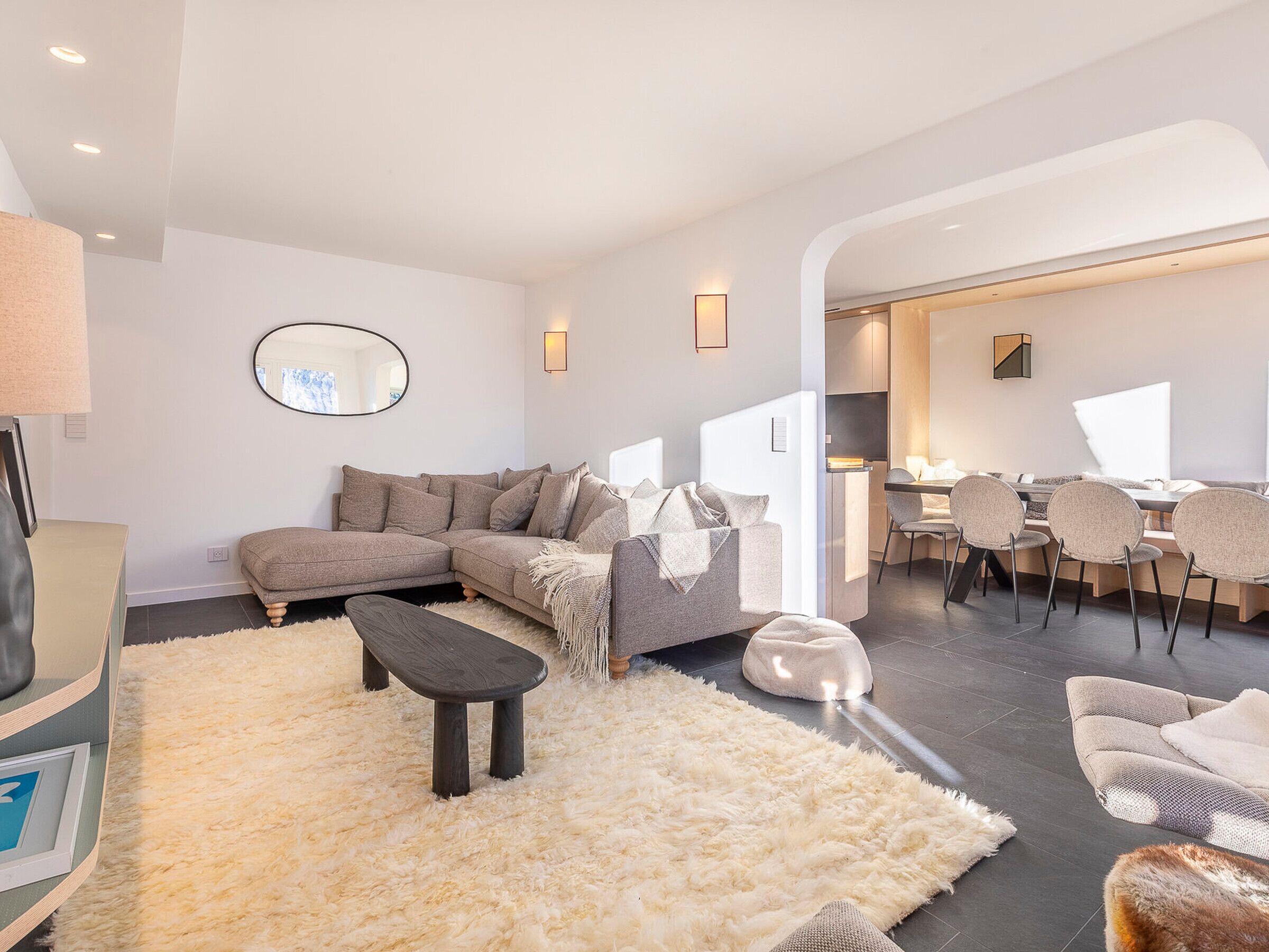 A modern living room with a sectional sofa, a small coffee table, and a fluffy rug. There is a dining area with a table and chairs visible through an open doorway.