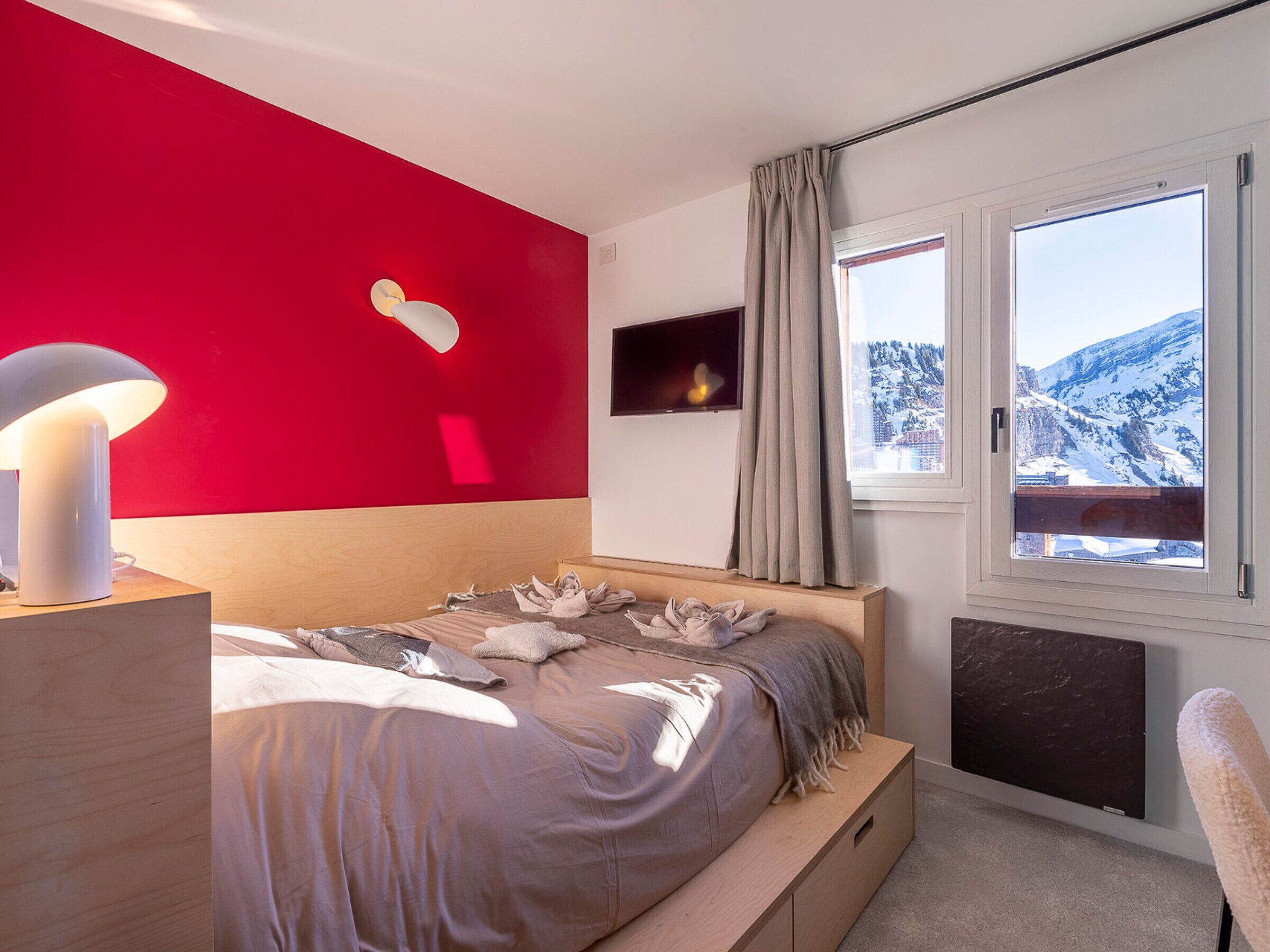 Ein Schlafzimmer mit Bett, Nachttischlampe, Fernseher und Fenster mit Blick auf einen verschneiten Berg.