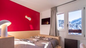 Ein Schlafzimmer mit Bett, Nachttischlampe, Fernseher und Fenster mit Blick auf einen verschneiten Berg.