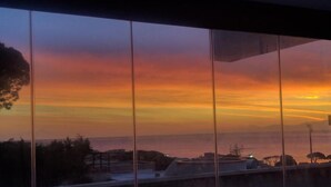 Une vue sur le coucher de soleil, prise depuis une fenêtre donnant sur une zone côtière avec des bâtiments et des arbres.