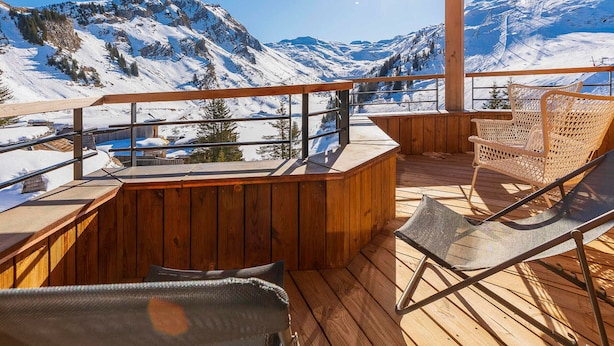 Une terrasse en bois offrant une vue sur des montagnes enneigées, un coin bar et deux fauteuils en osier.