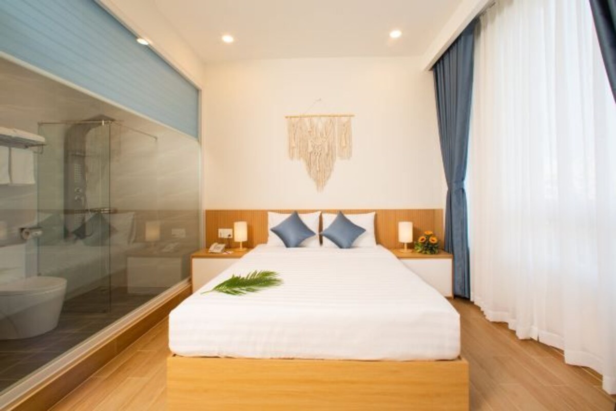 A modern hotel room with a large bed, a glass shower enclosure, wooden flooring, and a decorative wall hanging.