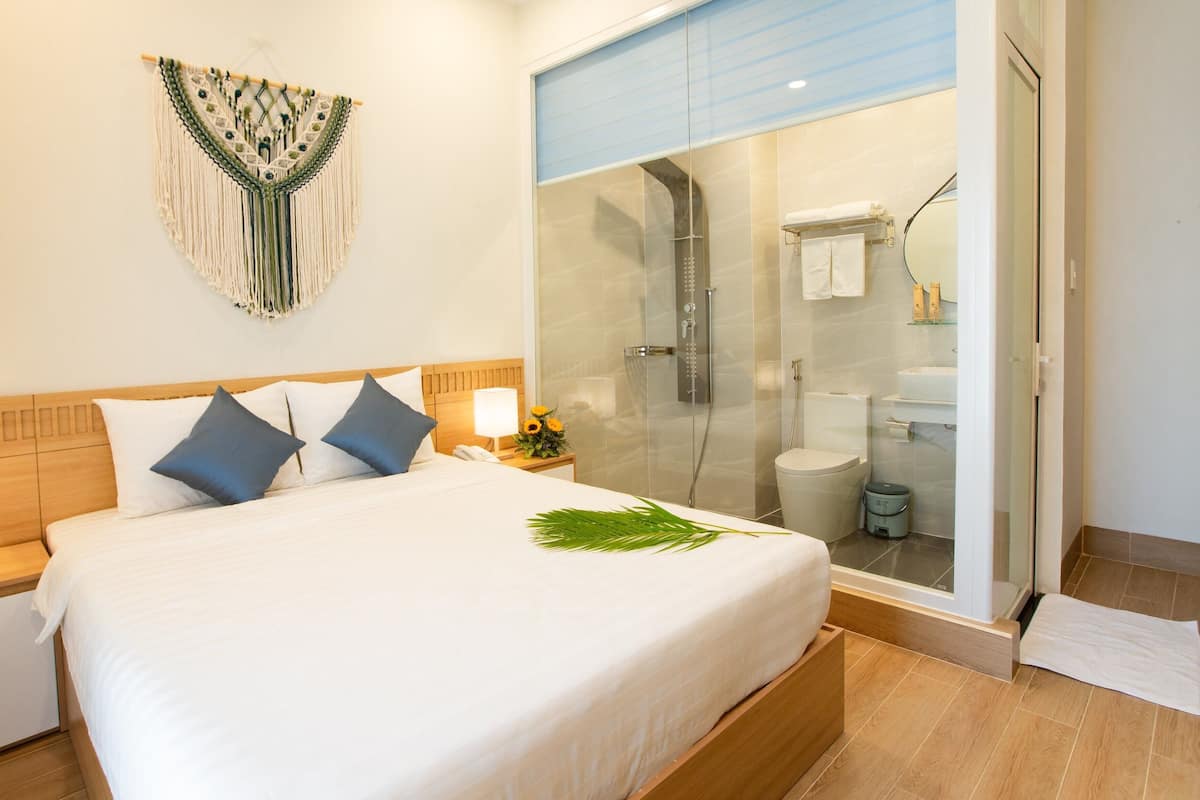 A hotel room with a bed, bedside lamps, a macrame wall hanging, and a glass-enclosed shower area.