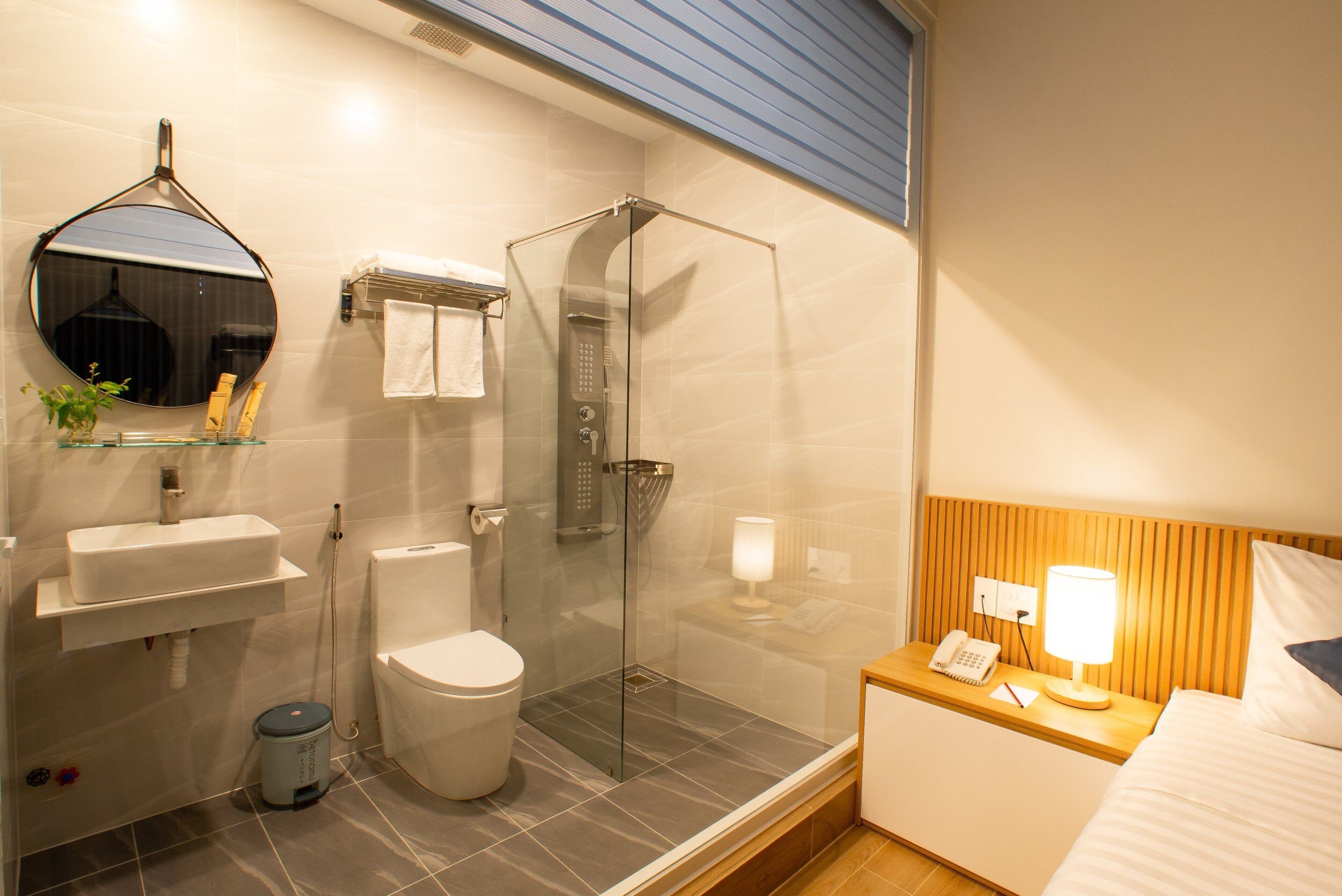 Un baño moderno con ducha de vidrio, inodoro, lavamanos y mesita de noche con lámpara.
