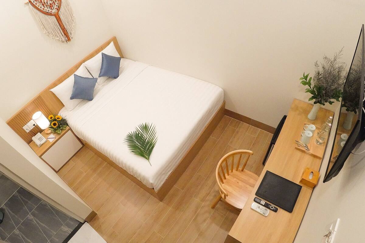 A compact hotel room with a bed, a wooden nightstand, a desk with a chair, a small table, and a TV.