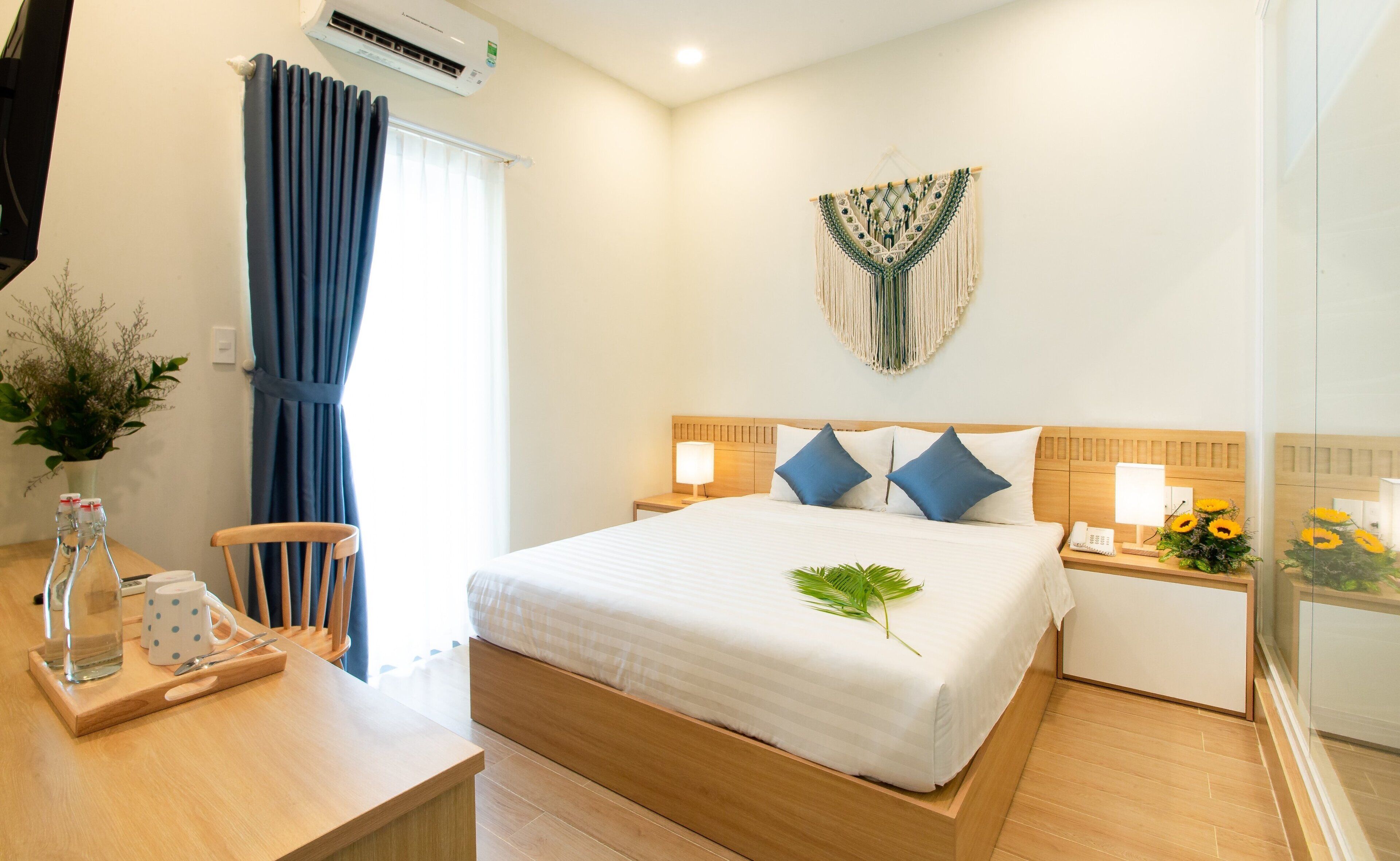 A hotel room with a bed, bedside tables, a desk with a chair, a potted plant, and a wall-mounted air conditioner.