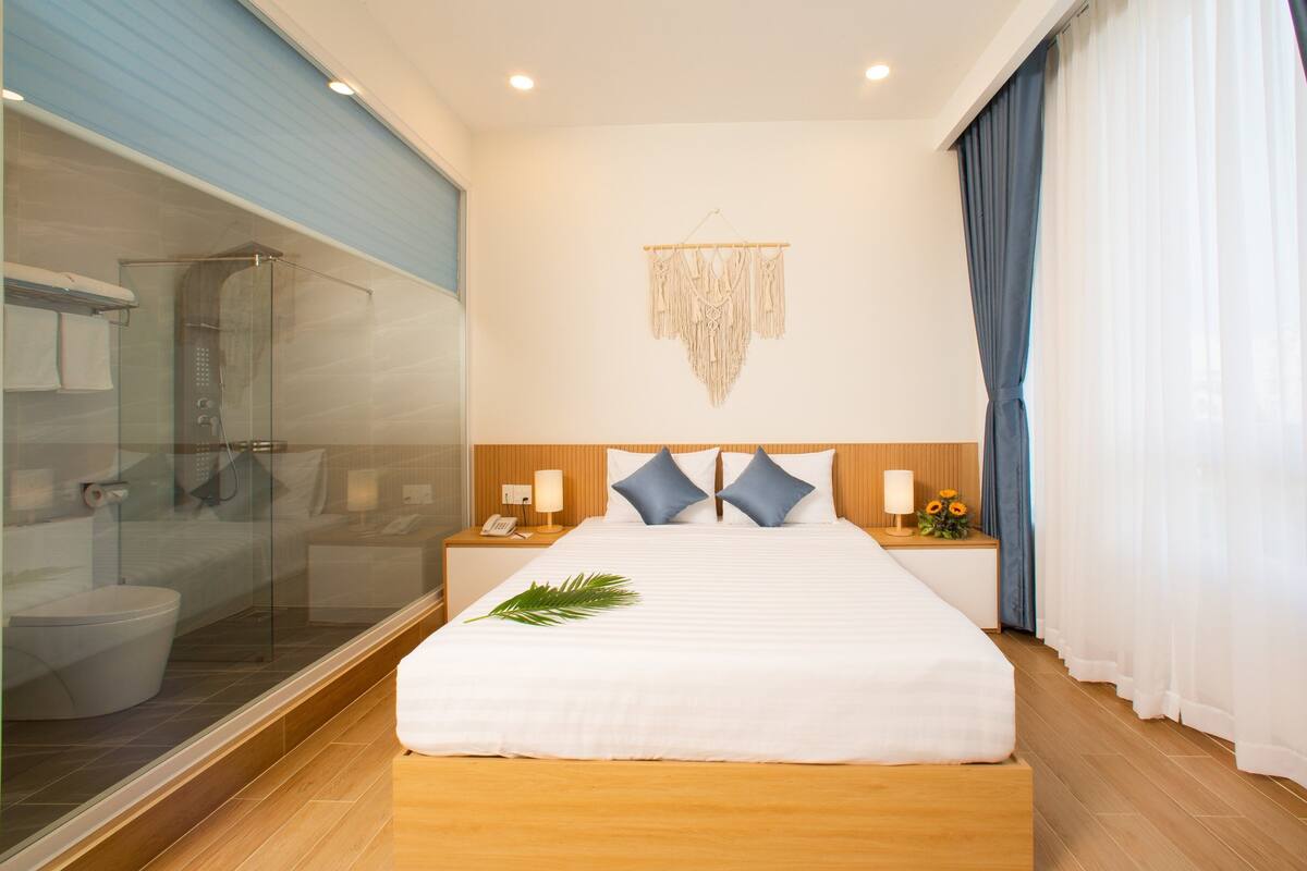 A hotel room with a bed, bedside tables, a wall-mounted macrame decoration, a shower area, and a view of the outside through a window.