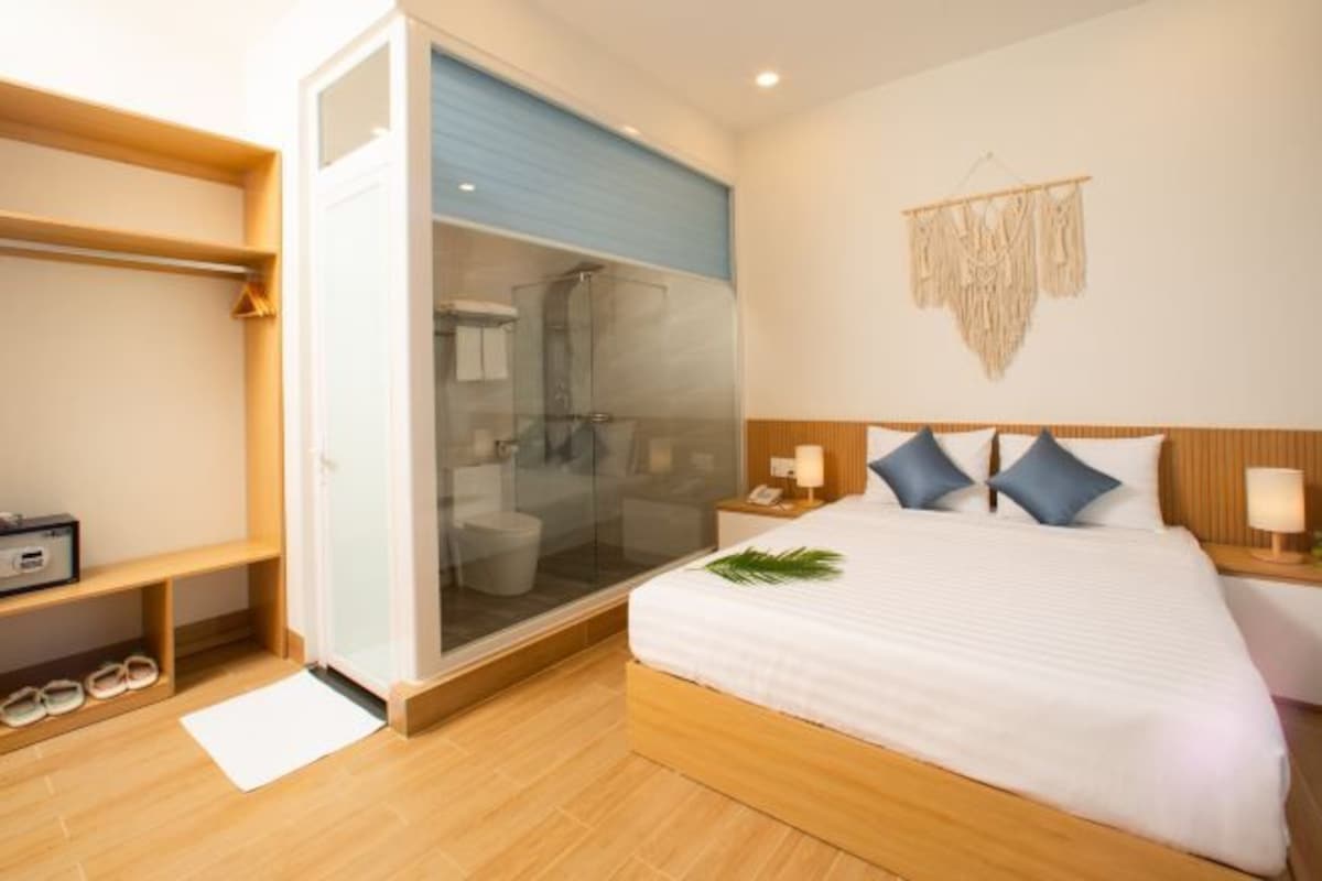 A modern hotel room with a wooden floor, a bed with white bedding and blue pillows, a glass shower enclosure, a wooden nightstand with a lamp, and a wall-mounted decor piece.