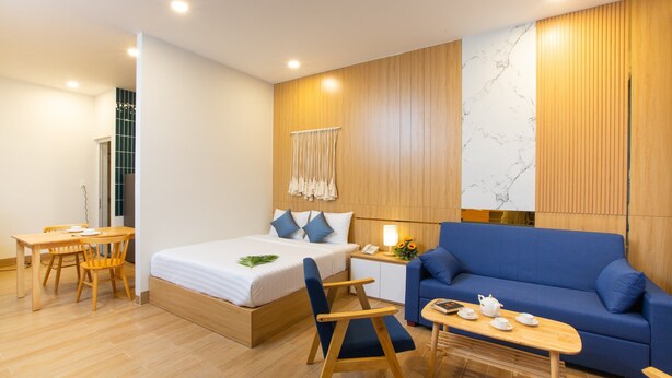 A modern hotel room with a bed, a blue sofa, a wooden table, and a small desk with a lamp and a phone. - Vy's House Phanthiet Hotel (Phu Thuy)