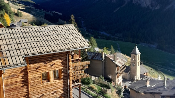 A rustic wooden building with a metal roof, a signboard with text, and a church with a bell tower. - Chalet Luxueux à 500 Mètres des Pistes Avec Terrasse Plein sud (Saint-Véran)
