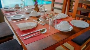 Een eettafel gedekt met witte borden, bestek en een glazen waterkan, met een zwembadzicht op de achtergrond.