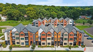 A multi-story residential building with a modern architectural design, featuring a mix of brick and concrete facades, and a parking area in front. - Tumbleweed -  Walk to Coffee + Dining + Garage (Shawnee)