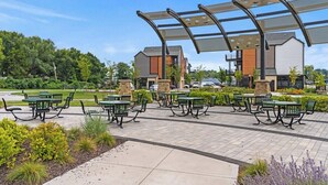 An outdoor patio area with multiple green benches and tables, surrounded by landscaped gardens and a modern building with a unique roof structure. - Tumbleweed -  Walk to Coffee + Dining + Garage (Shawnee)
