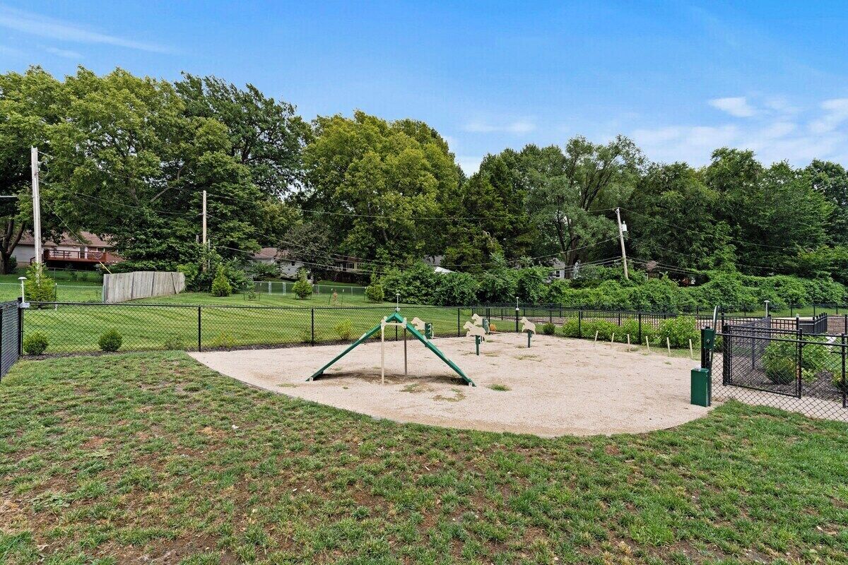 Un parque infantil con columpios y estructuras para escalar, rodeado por una zona vallada con árboles y un campo de césped.