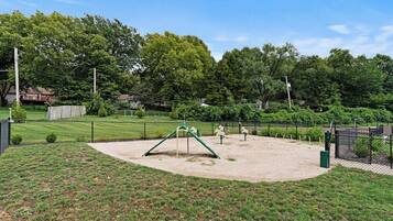 Un parque infantil con columpios y estructuras para escalar, rodeado por una zona vallada con árboles y un campo de césped.
