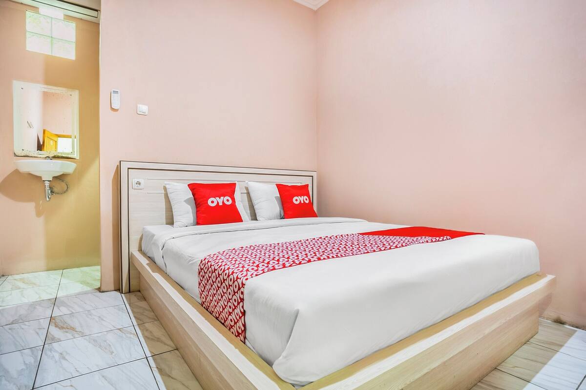 A neatly made bed with a white and red patterned bedspread, two pillows with "OYO" branding, a bedside table, and a mirror above a sink.