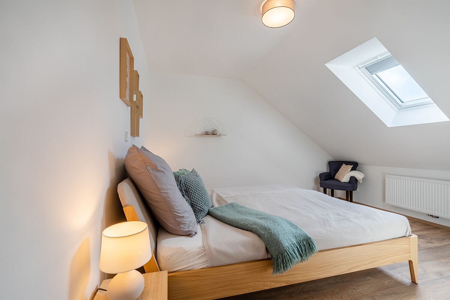 Un dormitorio con techo inclinado, tragaluces, cama con almohadas, mesita de noche con lámpara y una silla.