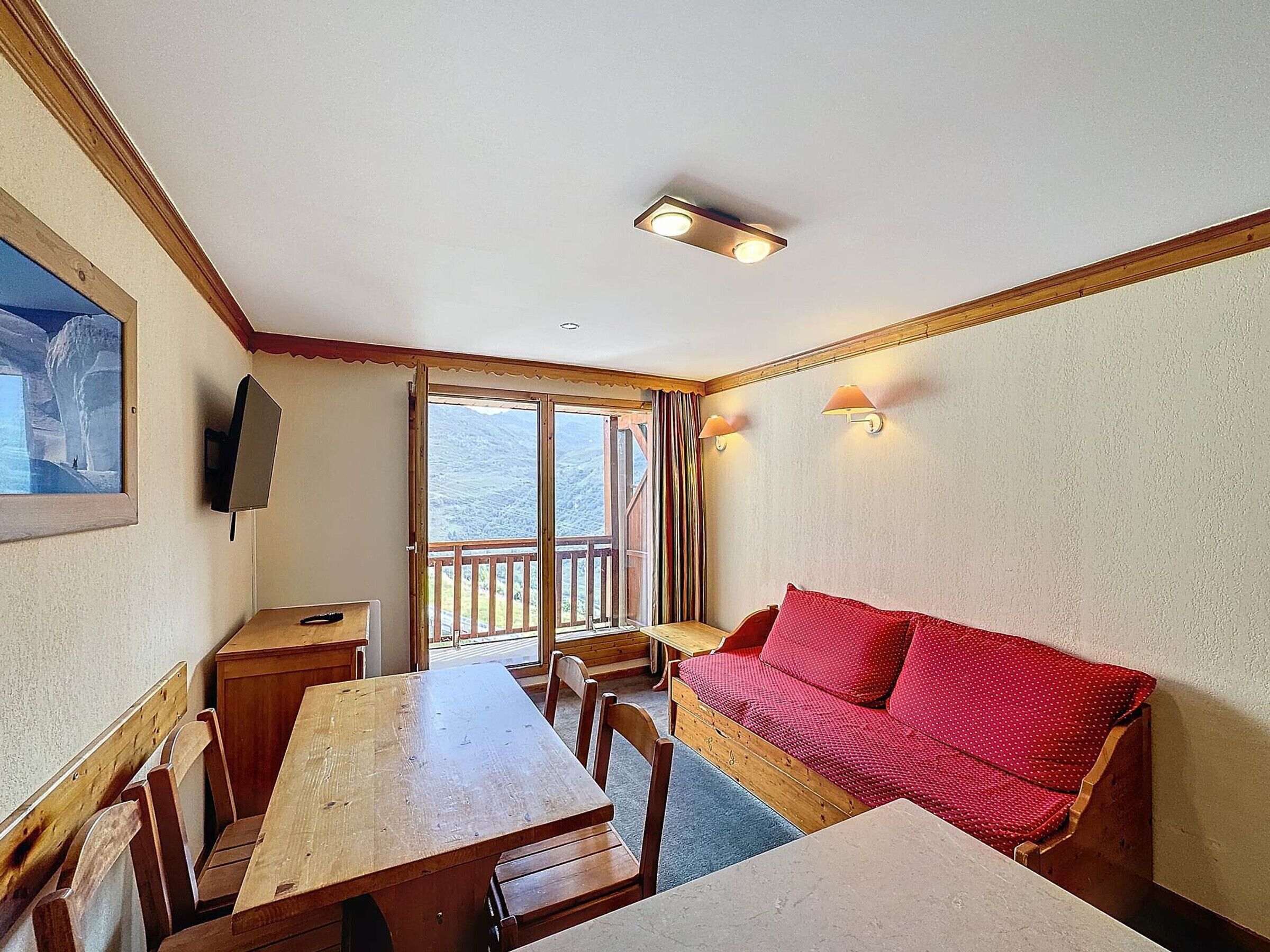 Una habitación con una mesa de madera, sillas, un sofá rojo, un televisor montado en la pared y un balcón con vistas a las montañas.