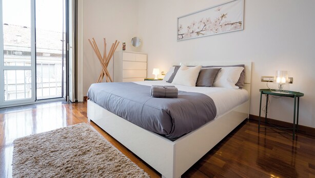 A bedroom with a large bed, a bedside table with a lamp, a small table with a lamp, a large window, and a painting on the wall. - notaMi - Cinque Vie Top Floor City Center (Milan)