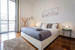 Un dormitorio con una cama grande, una mesita de noche con lámpara, una pequeña mesa con lámpara, un ventanal amplio y un cuadro en la pared.