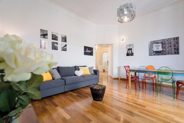 A modern living room with a grey sofa, a glass dining table, colorful chairs, and a wooden floor.