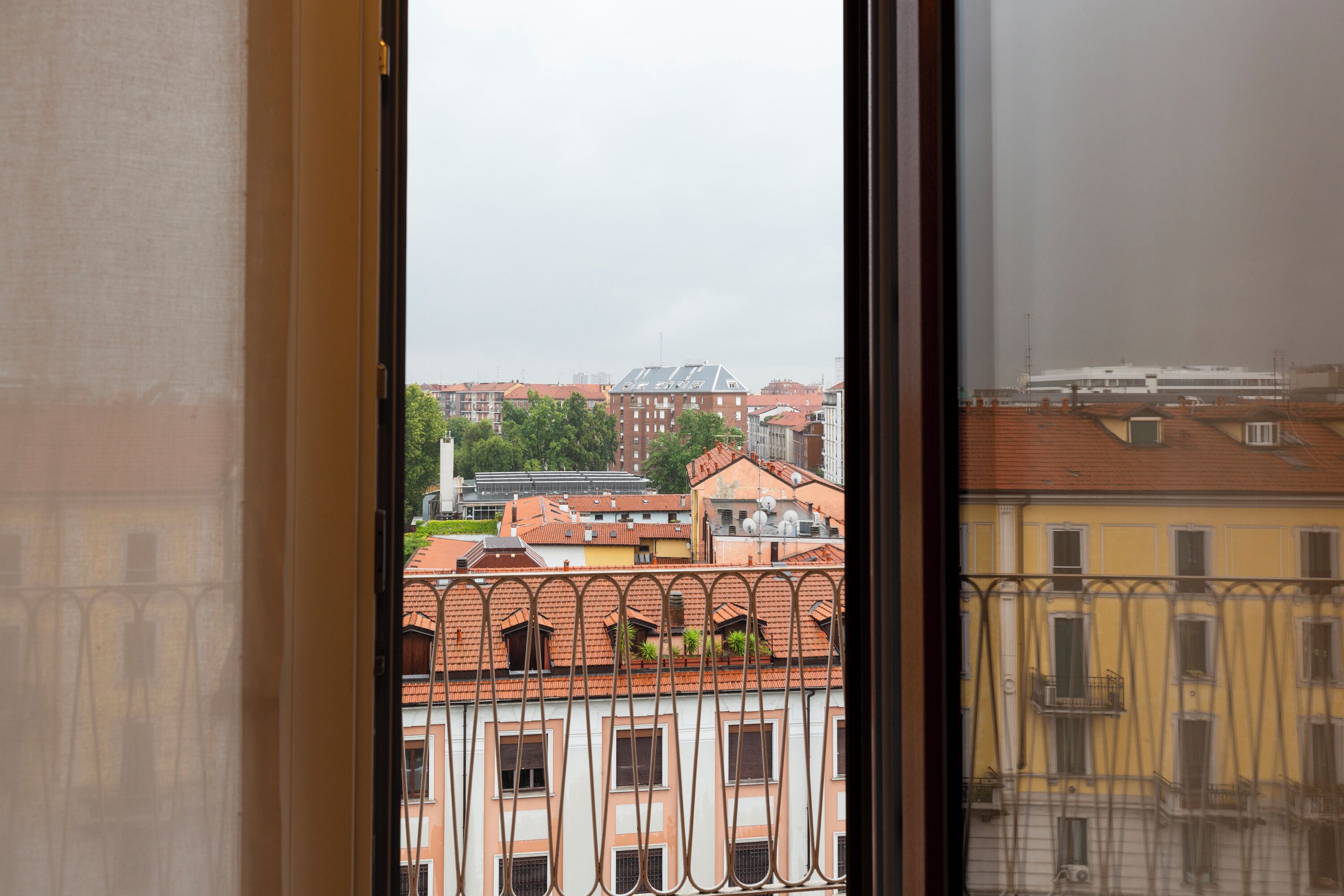 A view through a window with a metal railing, featuring buildings with red-tiled roofs and balconies.