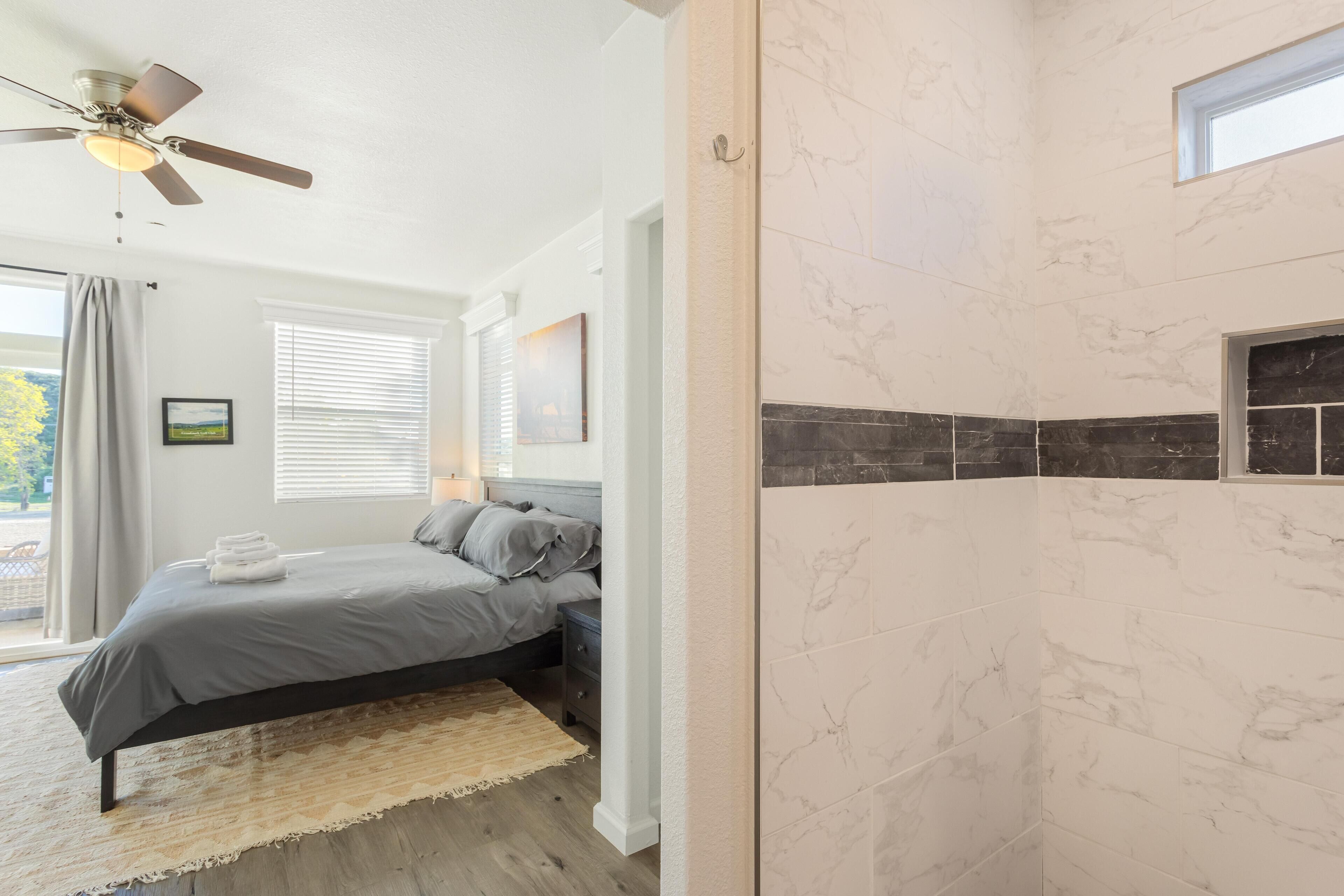 A bedroom with a bed, a ceiling fan, a window with blinds, and a bathroom with marble tiles.